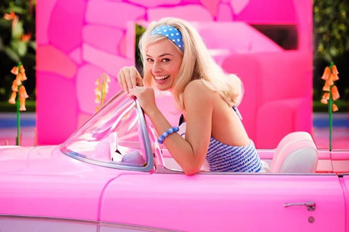 Imagem de cena do filme Barbie com personagem principal vestida com vestido, tiara e pulseira azuis, dentro de carro conversível cor de rosa