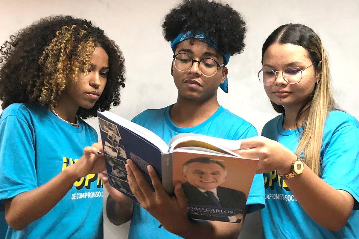 Imagem mostra três alunos do IJCPM segurando e lendo um livro sobre o empreendedor João Carlos Paes Mendonça