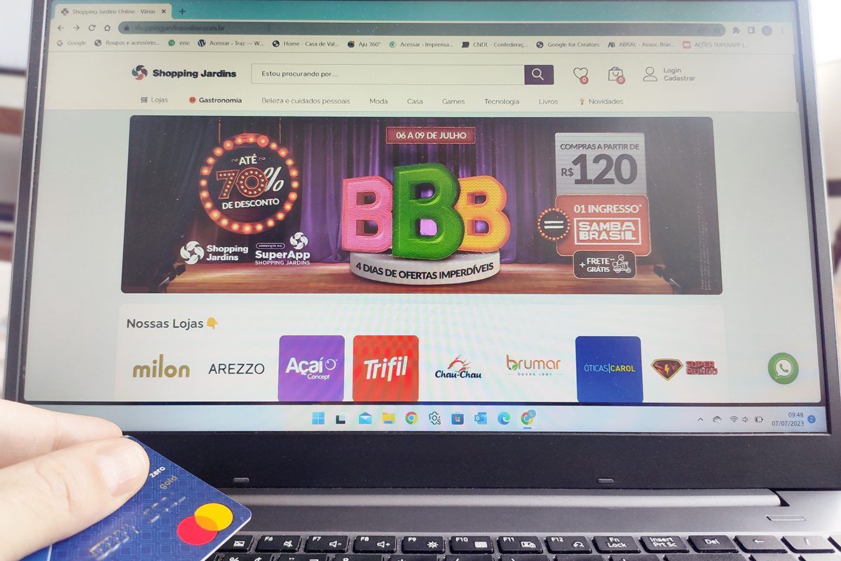 Imagem de usuário com cartão de crédito em mãos acessando o canal de compras online do Shopping Jardins pelo laptop. Na tela, há banner sobre a promoção compre e ganhe ingresso para o evento Samba Brasil Aracaju