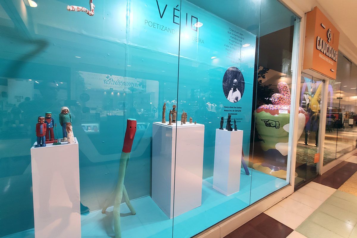 Imagem da vitrine do projeto Cajucidade no Shopping Jardins com a exposição Véio, Poetizando a Madeira em primeiro plano