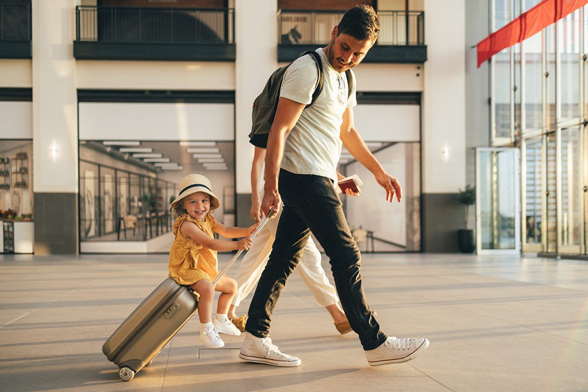 Imagem de família no aeroporto com pai empurrando a mala com filha sentada encima