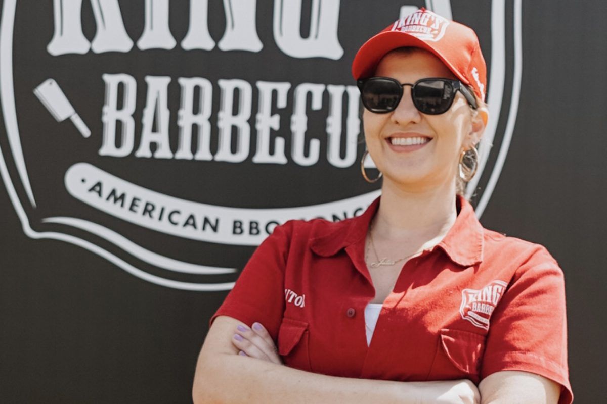 Imagem da chef Júlia Carvalho que apresentará aula-show gratuita de churrasco. A chef está de braços cruzados, vestindo camisa e boné vermelhos e óculos de sol