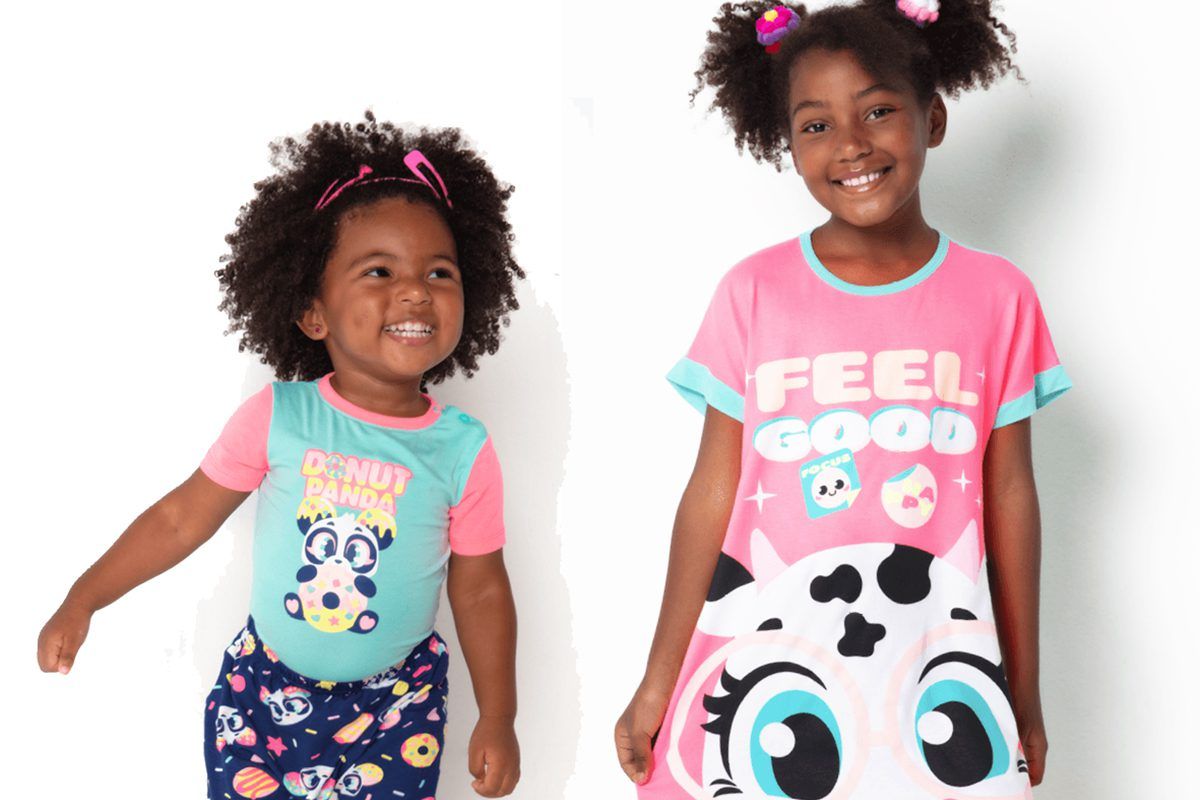 Imagem de duas crianças pretas sorrindo. Uma veste conjunto e outra vestido Puket, coloridos e com estampa de unicórnio